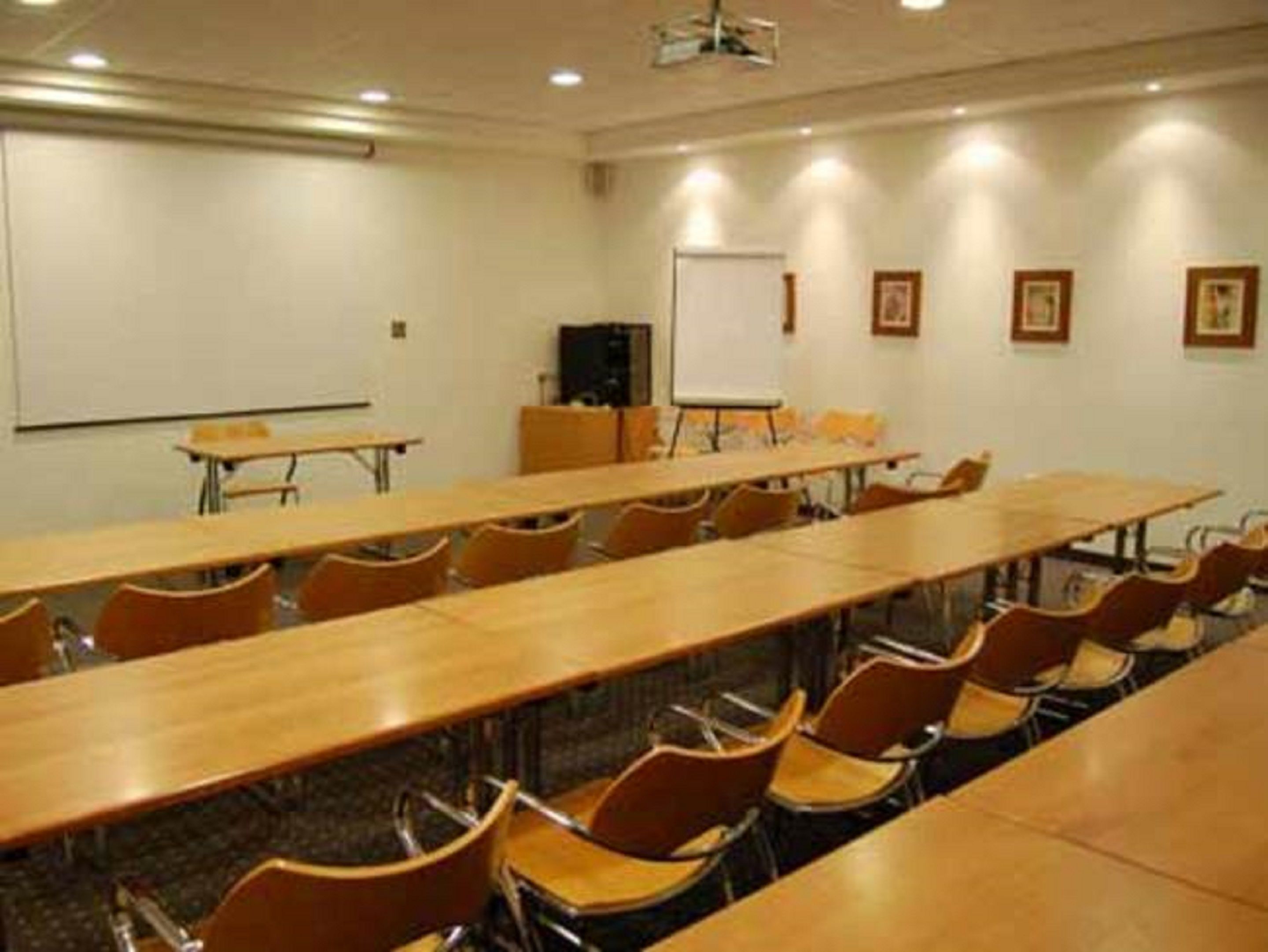 Een vergaderzaal met lange tafels en stoelen, een whiteboard en ingelijste foto’s aan de muur.