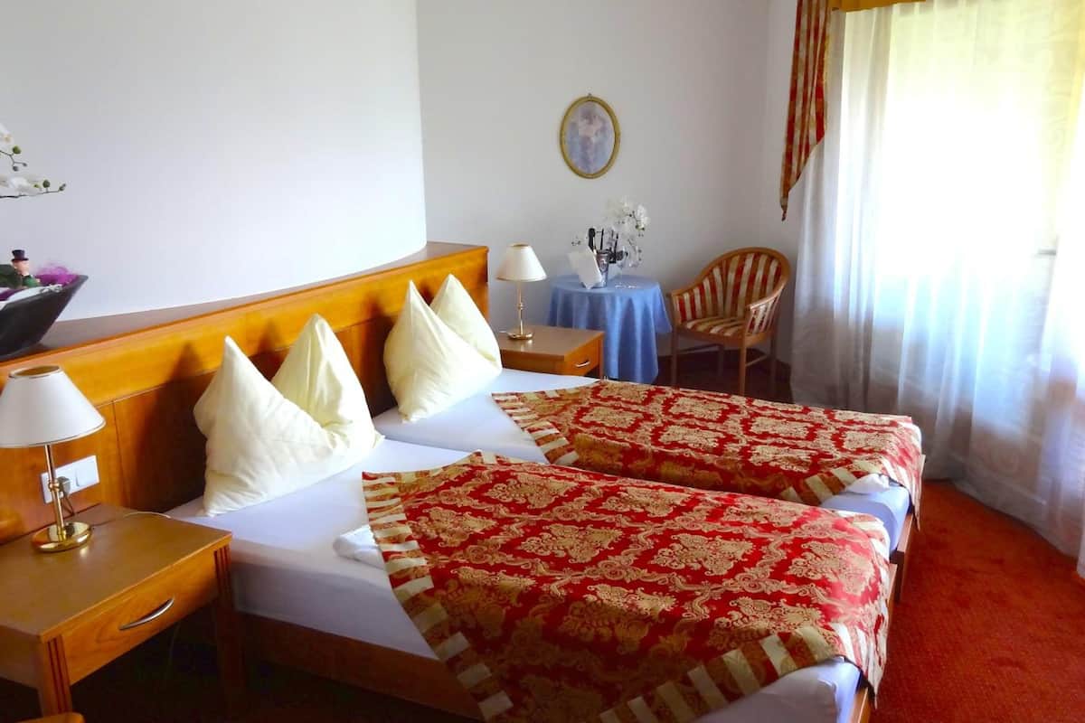 A hotel room with two beds, a bedside table with a lamp, a small table with a vase of flowers, a chair, and a window with curtains.