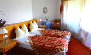 Ein Hotelzimmer mit zwei Betten, einem Nachttisch mit Lampe, einem kleinen Tisch mit Blumenvase, einem Sessel und einem Fenster mit Vorhängen.