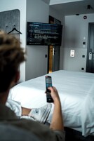Uma pessoa segurando um controle remoto num quarto de hotel com uma cama, uma televisão montada exibindo um menu, e uma porta com fechadura e leitor de cartão-chave.