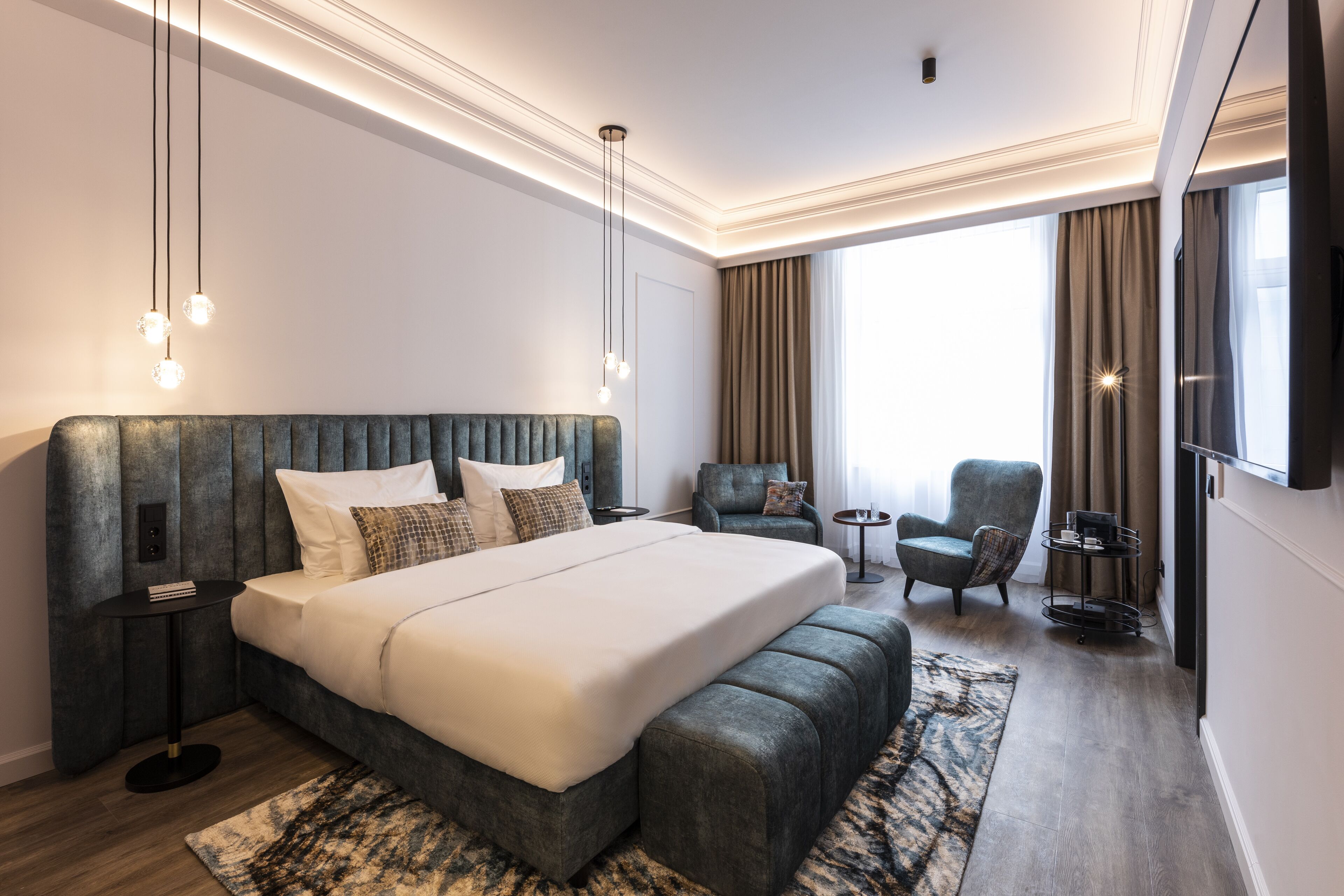 A modern hotel room with a large bed, a seating area with a chair and a small table, and a TV mounted on the wall.