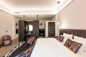 Quarto de hotel moderno com uma cama grande, área de estar com tapete estampado, um espelho redondo pequeno e um guarda-roupa de madeira.