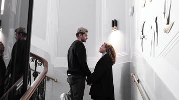 Ein Mann und eine Frau stehen auf einer Treppe in einer Hotelhalle, mit einer weißen Wand und einem Geländer.