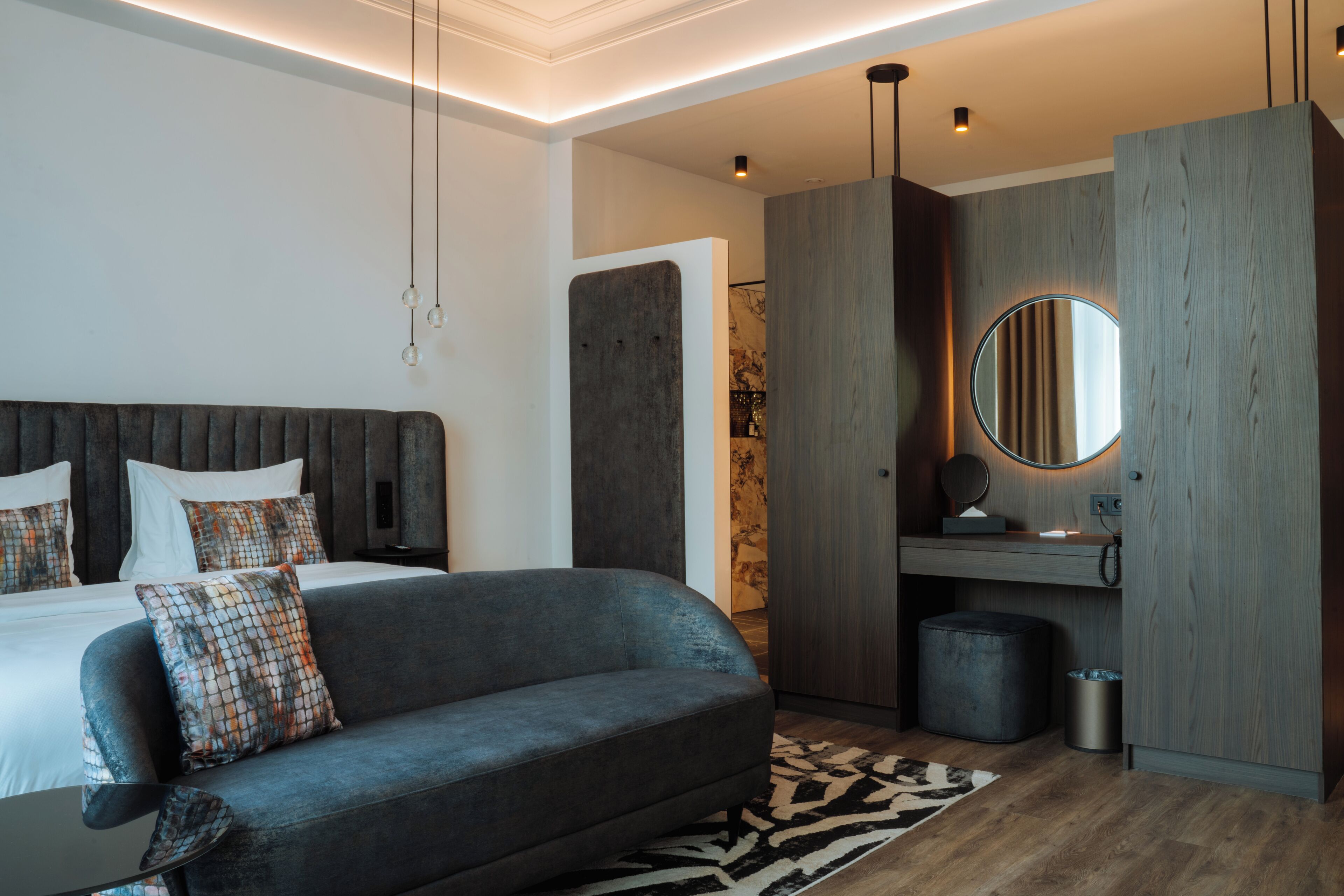 A modern bedroom with a bed, a grey sofa, a round mirror, and a wooden wardrobe.