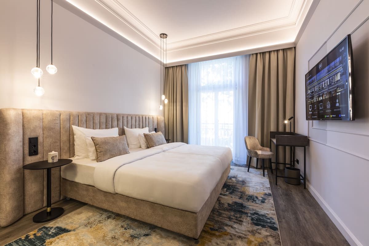 A modern hotel room with a large bed, a side table, a chair, a TV, and a large window with curtains.