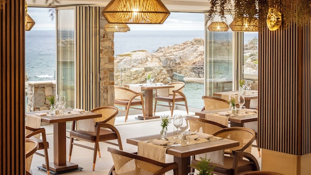 Un restaurante con terraza al aire libre y vistas al mar, con mesas y sillas de madera y grandes ventanales.