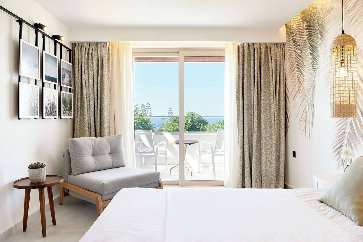 A modern hotel room with a bed, a seating area, a balcony with a table and chairs, and a view of the sea.