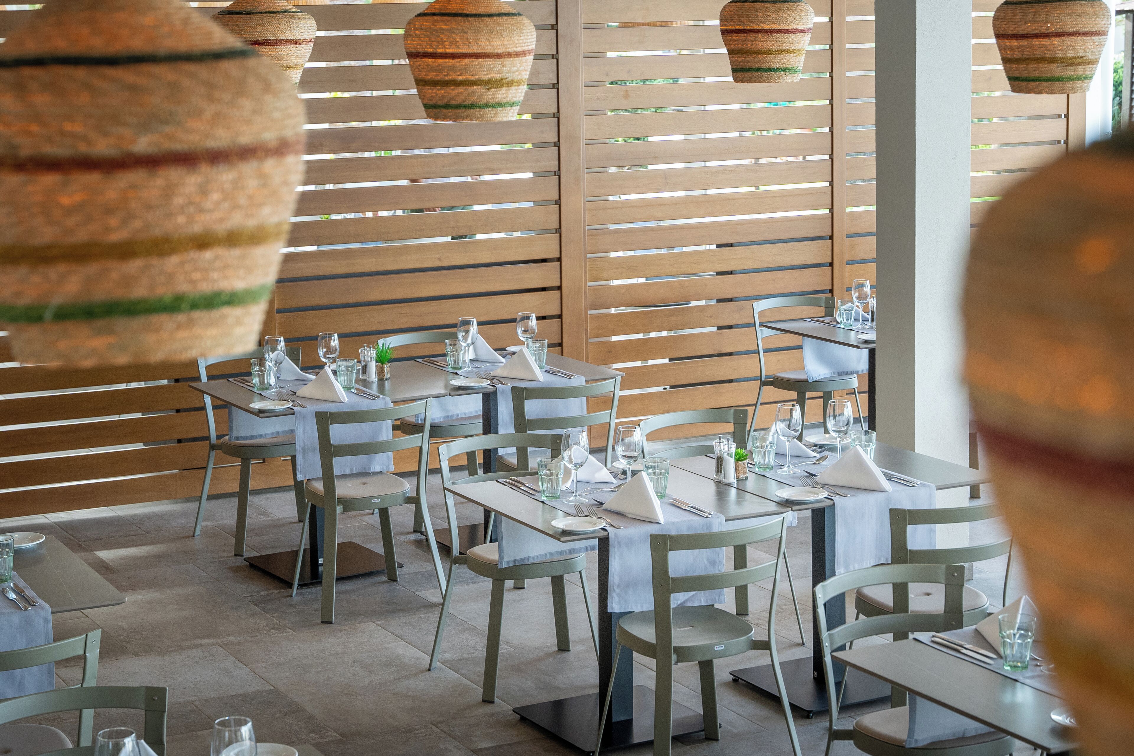 A restaurant with tables set for dining, hanging woven baskets, and a slatted wooden wall.