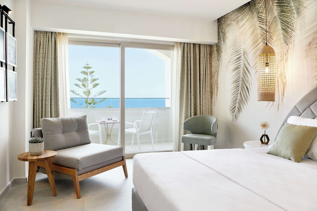 A hotel room with a bed, a sofa, a chair, a small table, and a view of the sea through a large window.