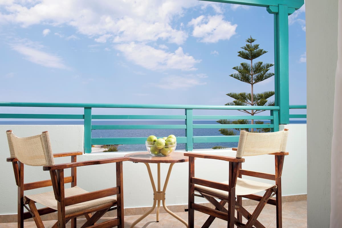 A balcony with two wooden chairs and a small table, overlooking a turquoise railing and a clear sky.