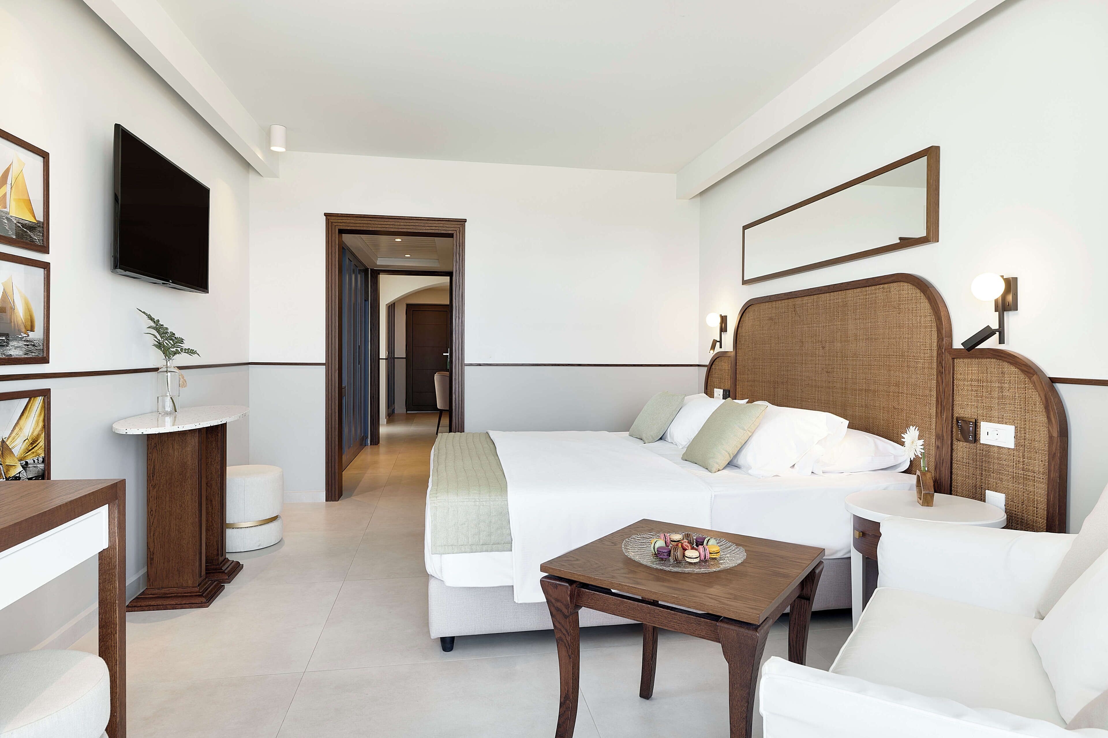Habitación de hotel con cama, mesita de noche con un objeto decorativo, una mesa redonda, una silla, televisor montado en la pared y un espejo sobre la cama.