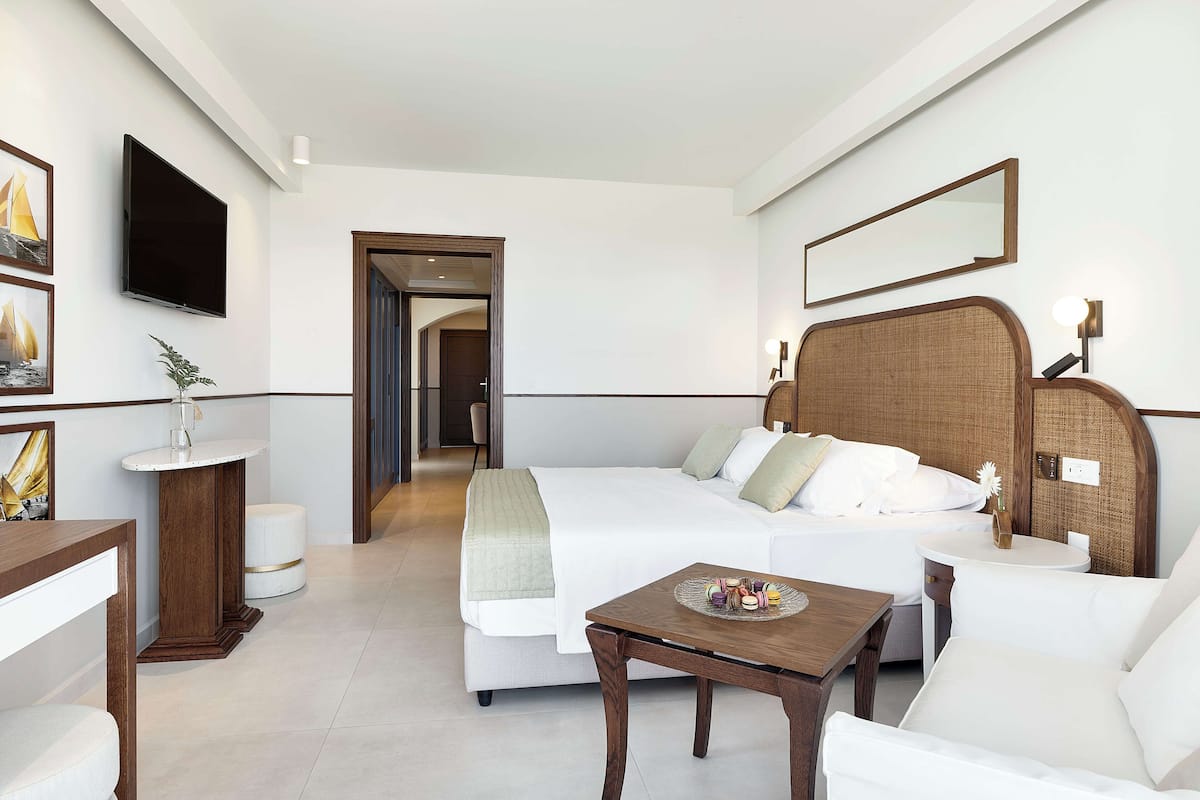 Habitación de hotel con cama, mesita de noche con un objeto decorativo, una mesa redonda, una silla, televisor montado en la pared y un espejo sobre la cama.