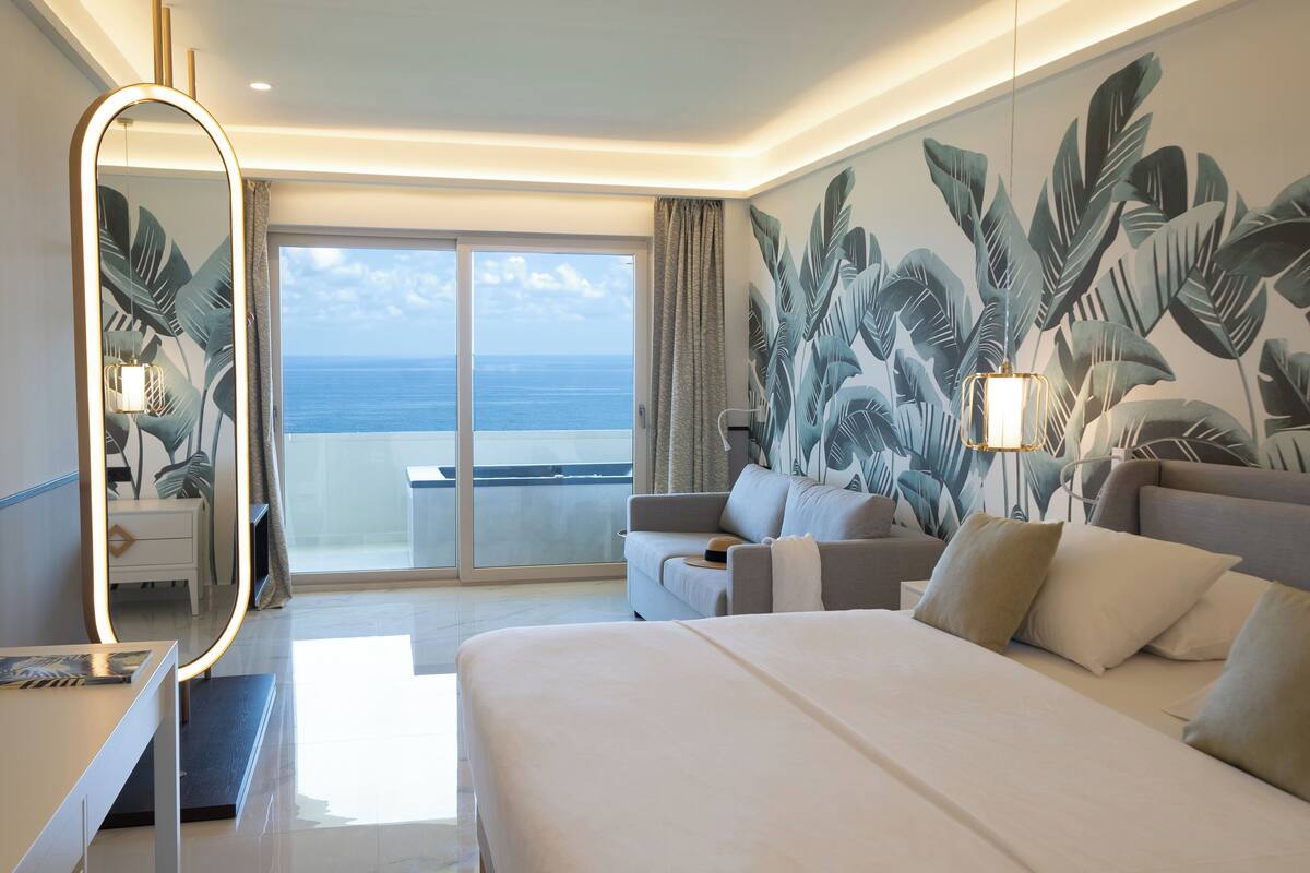 A hotel room with a large bed, a sitting area with a sofa and armchairs, a large mirror, and a view of the ocean through a sliding door.