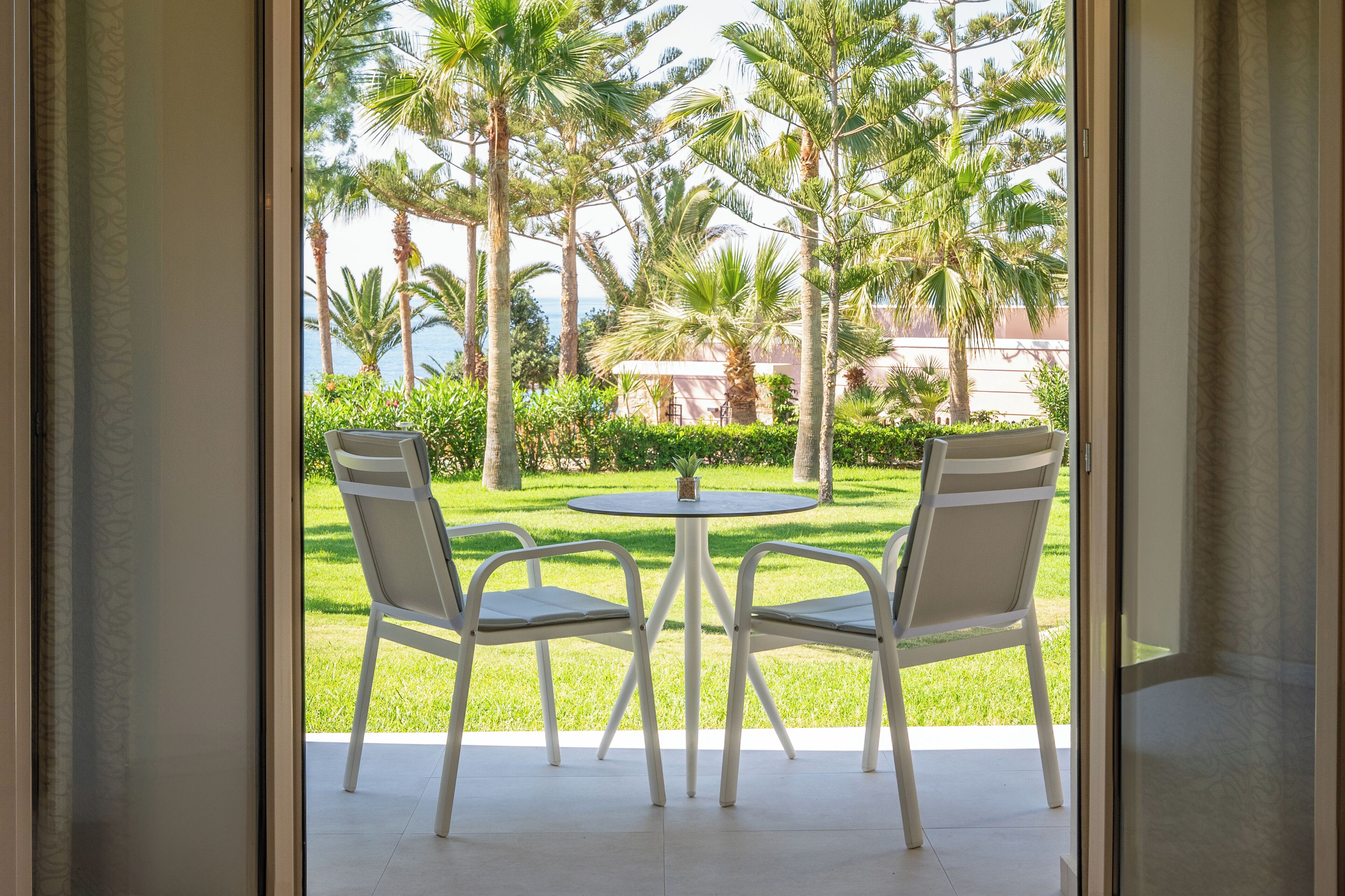 A view from a room with a glass door to an outdoor patio with a round table and two chairs, surrounded by palm trees and a well-maintained lawn.