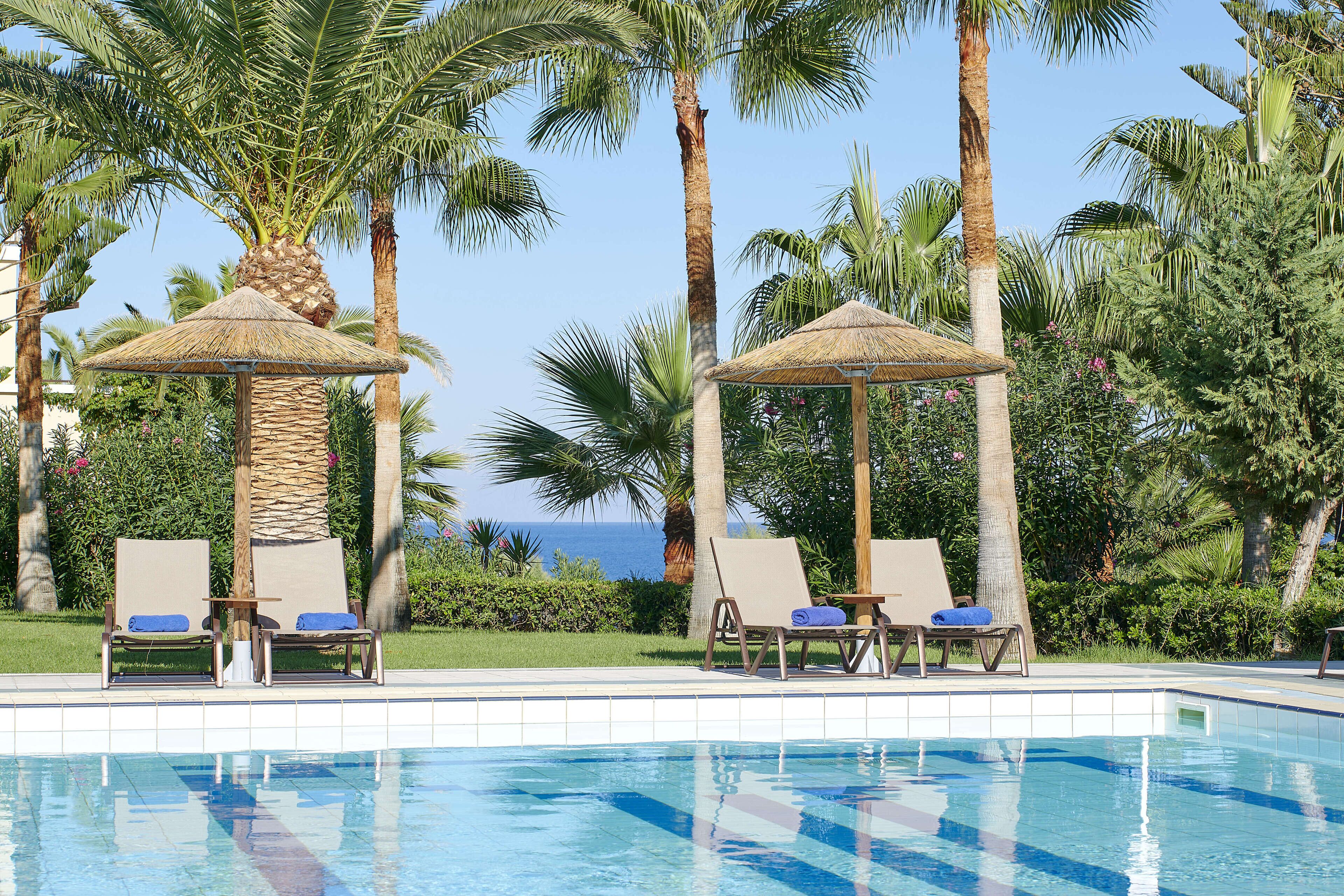 Un espace piscine agrémenté de chaises longues et de parasols, entouré de palmiers et offrant une vue sur l’océan.