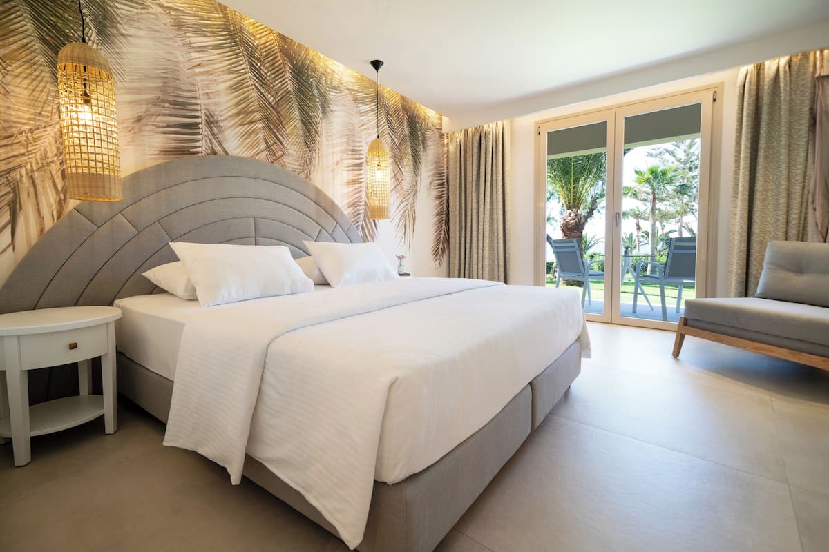 A modern bedroom with a large bed, a nightstand, a sofa, and a view of palm trees through the glass door.