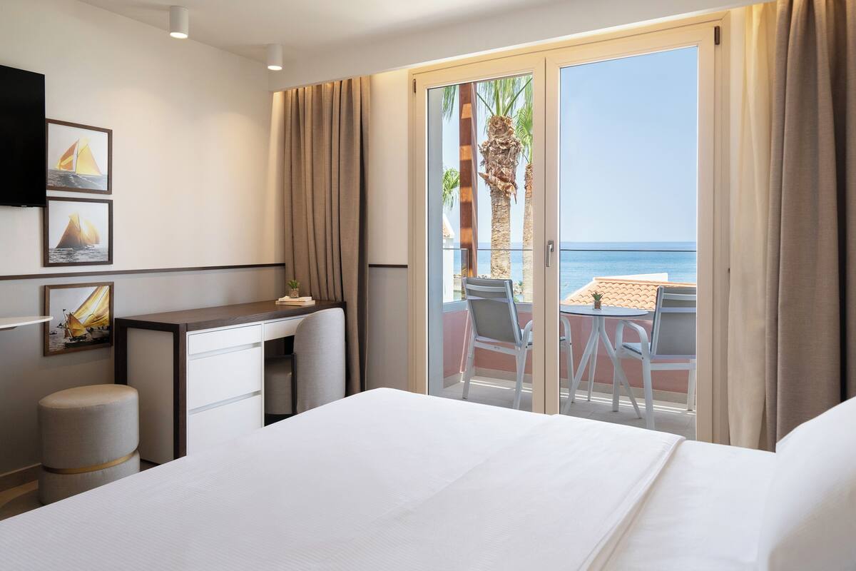 A hotel room with a bed, a desk, a chair, and a balcony with a view of the sea.