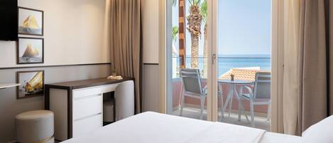 A hotel room with a bed, a desk, a chair, and a balcony with a view of the sea.