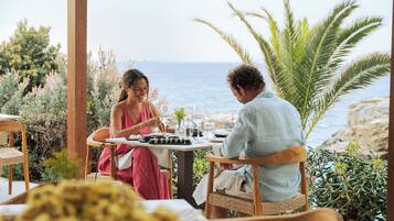 Una pareja cenando en una mesa con vista al mar, rodeada de plantas y muebles de madera.