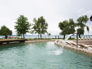 A swimming pool with a water feature, surrounded by lounge chairs and trees, with a view of the sea in the background. - Prémium Hotel Panoráma (Siofok)