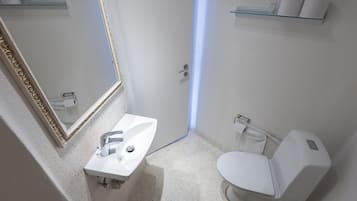 Un baño moderno con un lavabo blanco, espejo y retrete. Hay un estante de cristal sobre el retrete y un dispensador de toallas a la derecha.