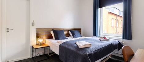 A hotel room with a bed, a bedside table with a lamp, a chair, and a window with curtains.