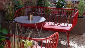 Un espace extérieur aménagé avec des sièges en métal rouge, comprenant une table ronde et des chaises, le tout entouré de plantes en pot.