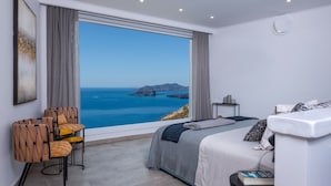 A modern bedroom with a large window offering a view of the sea and a mountainous landscape. - Athermi Suites - Adults Only (Santorini)