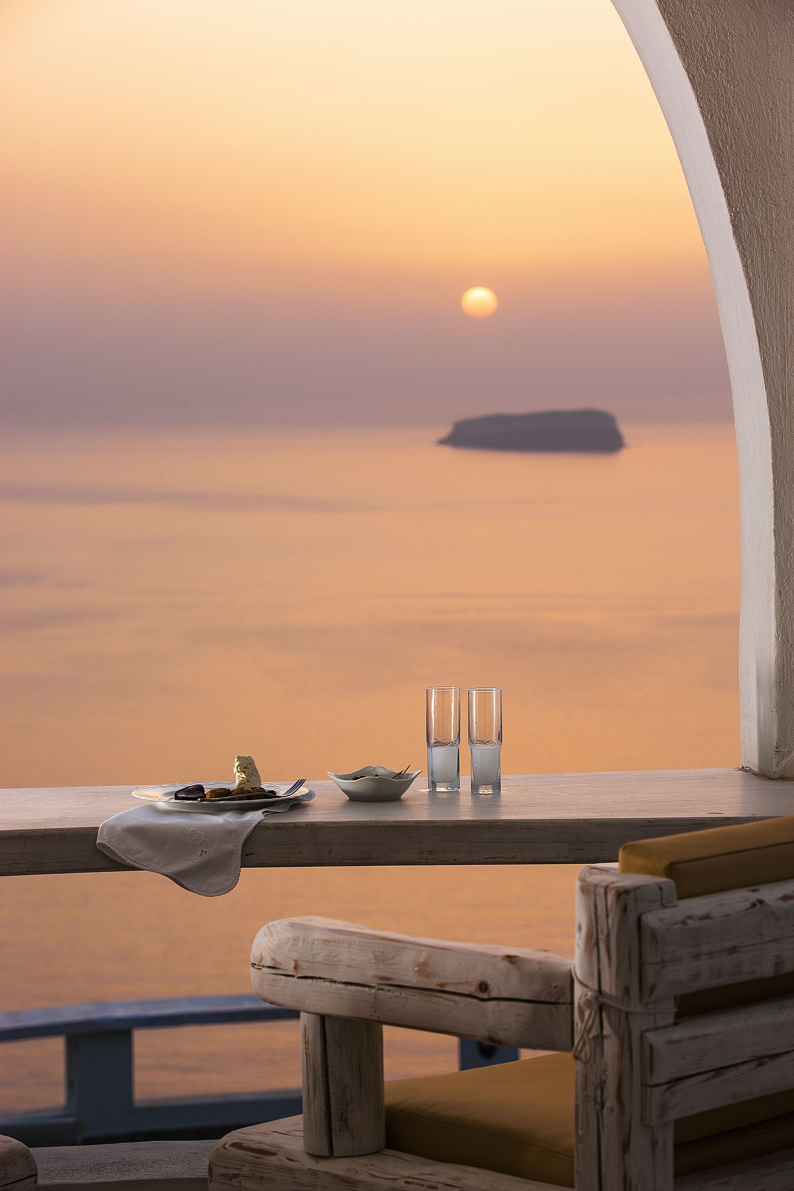 A table with a plate, two glasses, and a napkin, overlooking a sunset over the sea.