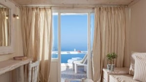 A room with a view of the sea, a chair, a small table with a plant, and a window with curtains. - Athermi Suites - Adults Only (Santorini)