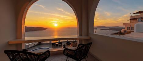 A balcony with two chairs facing a sunset view over the sea, a pool, and a modern building in the background.