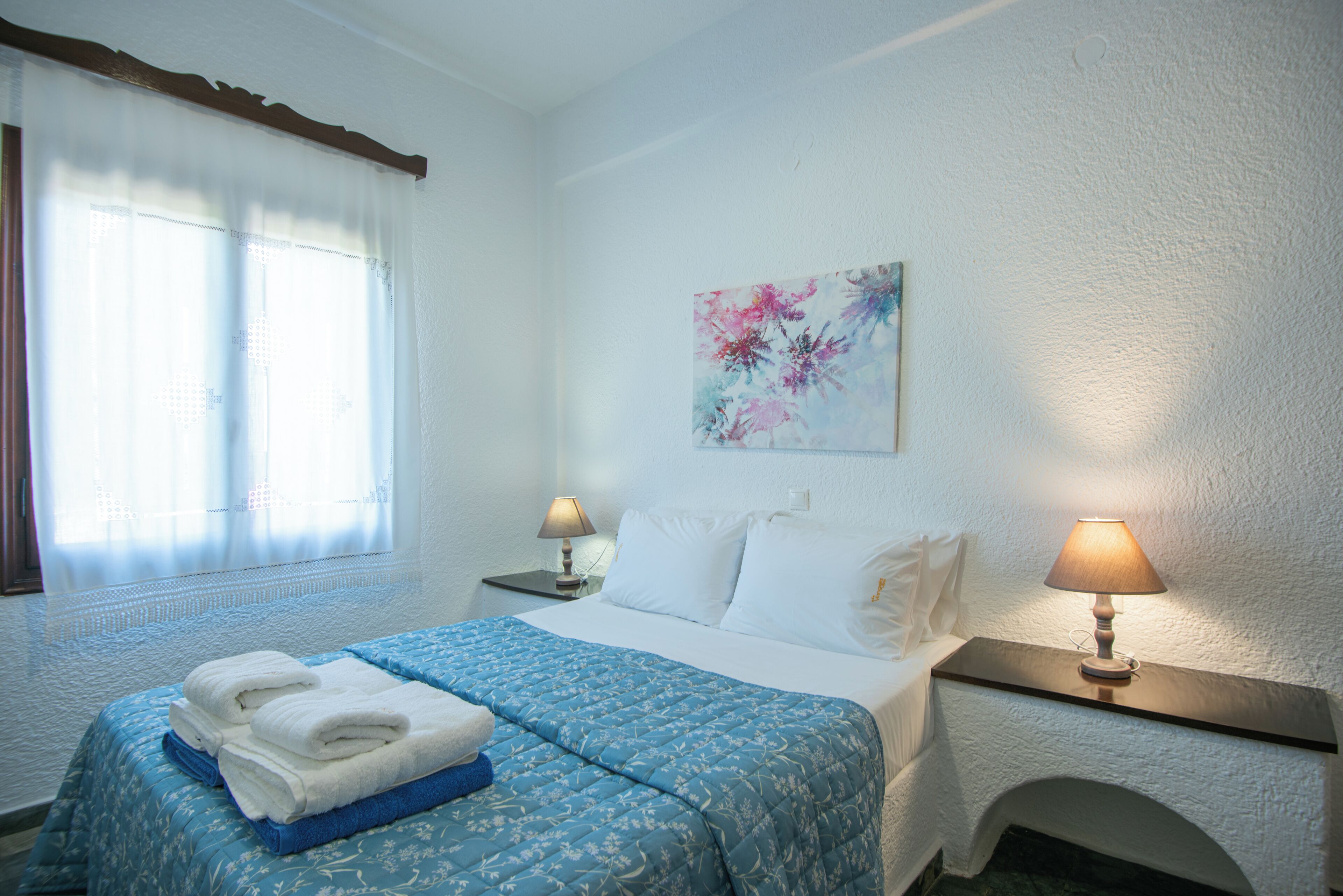 A hotel room with a bed, two bedside lamps, a painting on the wall, and a window with curtains.