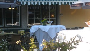 Een terras met eettafels, gedekt met witte tafelkleden, potplanten en een raam met uitzicht op de straat.