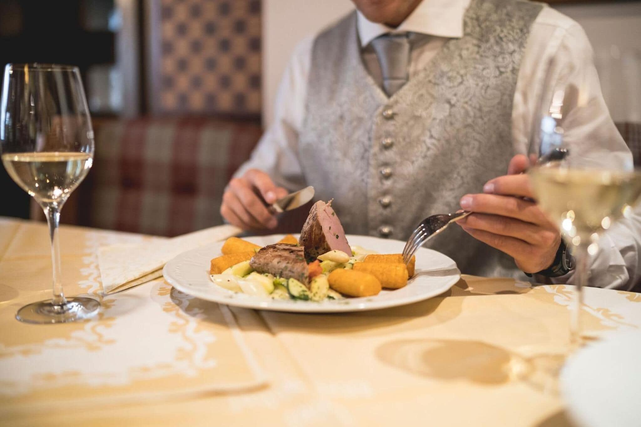 A dining table with a plate of food, a wine glass, and a person in a vest.