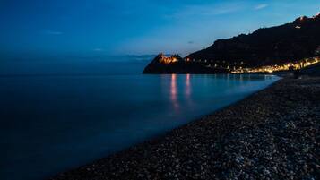 A pebbled beach at dusk with a rocky outcrop in the distance and lights along the shoreline.