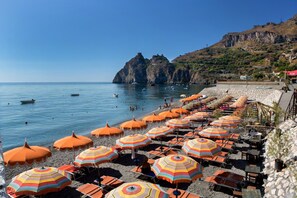 Una playa con hileras de sombrillas naranjas y tumbonas, un litoral rocoso y un acantilado destacado al fondo.