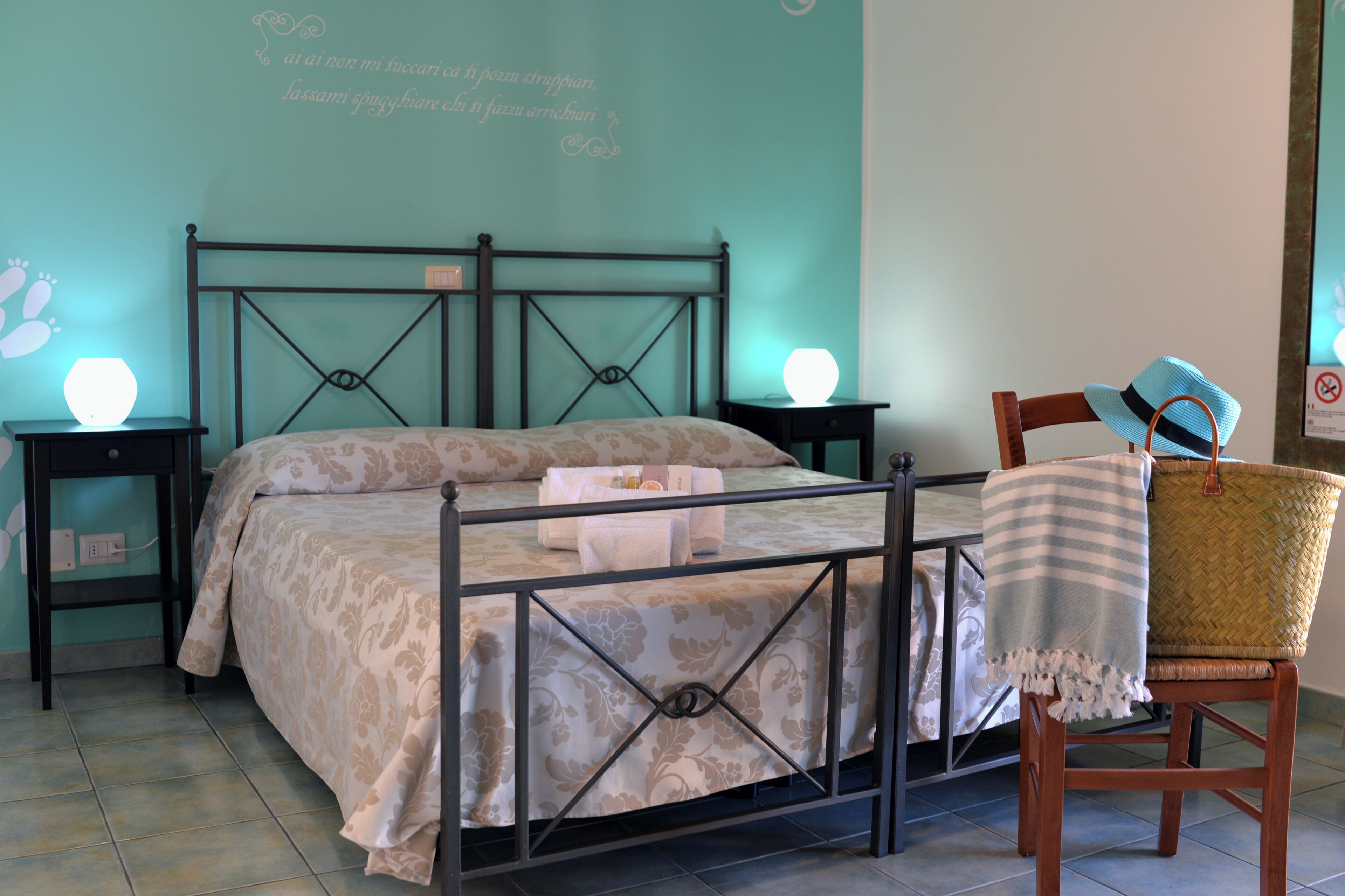 Un letto rifatto con una coperta a fantasia e cuscini, due comodini con lampade, una sedia in vimini con un cappello, e una parete con scritte decorative.