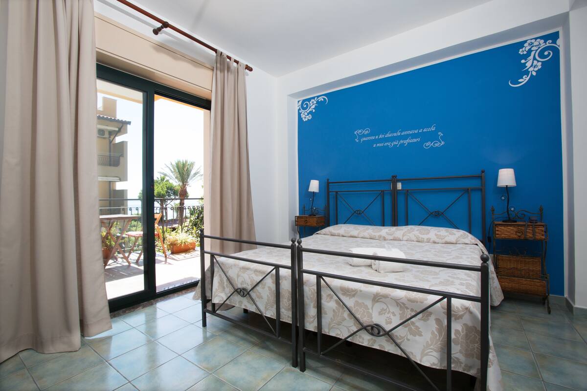 Ein Schlafzimmer mit Bett, Balkonblick und blauer Wand mit dekorativem Text.