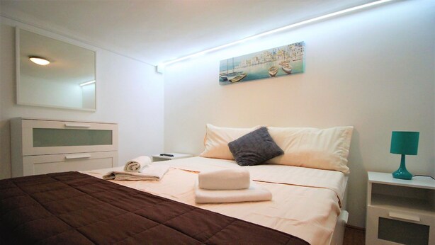 A neatly made bed with a brown bedspread, a white pillow, and a folded towel. There is a white bedside table with a teal lamp, a white dresser with a mirror, and a painting of boats on the wall.