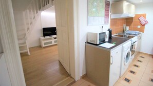 A compact kitchen with a microwave, washing machine, and a small sink. A television is placed on a white stand in the adjacent room.