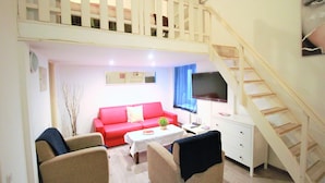 A living room with a red sofa, beige armchairs, a white coffee table, and a TV on the wall. A staircase with a wooden railing is visible to the right.