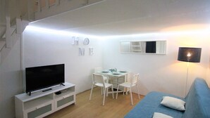 A compact living space with a TV on a white stand, a dining table with chairs, a sofa with cushions, and a staircase in the background.