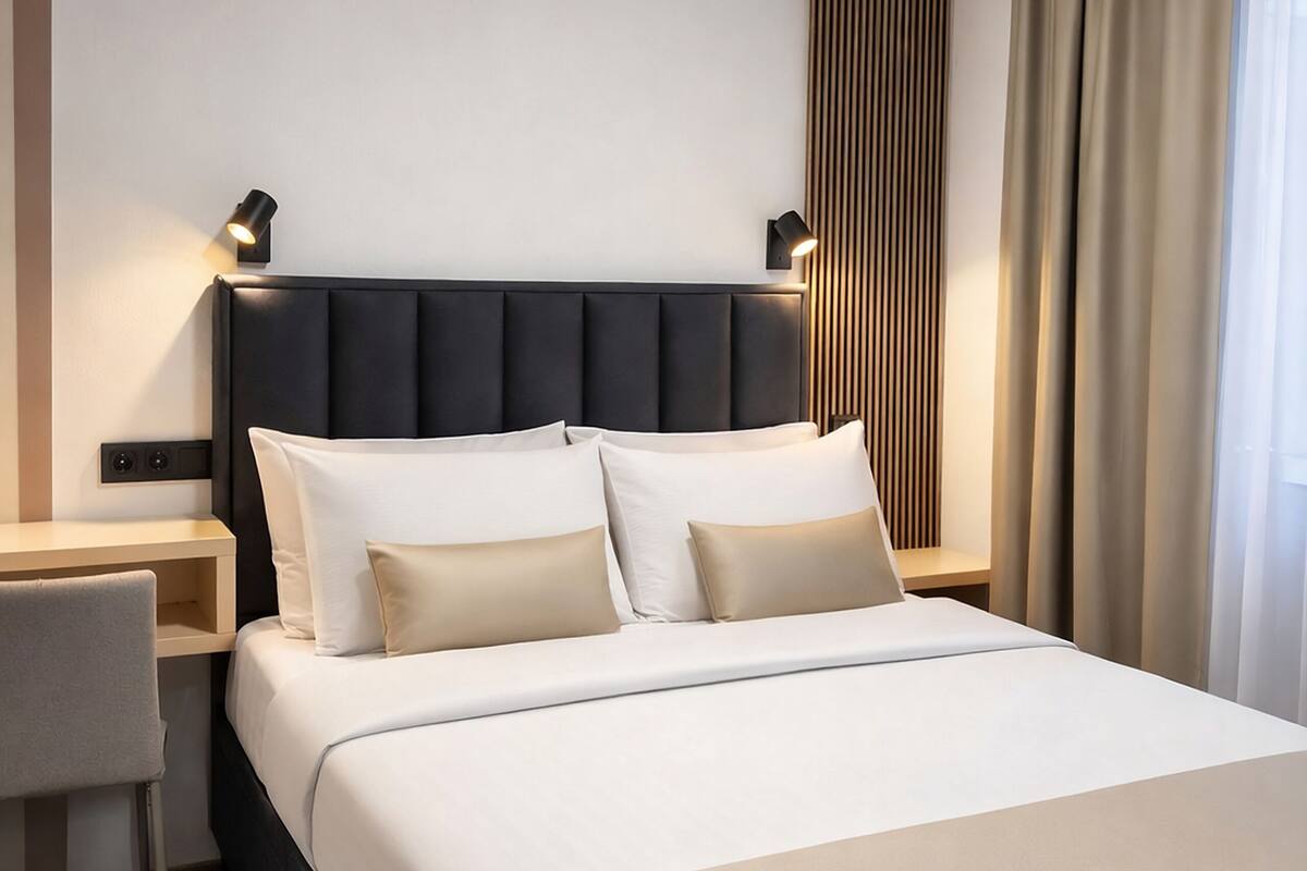 Ein modernes Hotelzimmer mit einem ordentlich bezogenen Bett, einem Nachttisch, einem Sessel und einem großen Fenster mit Vorhängen.