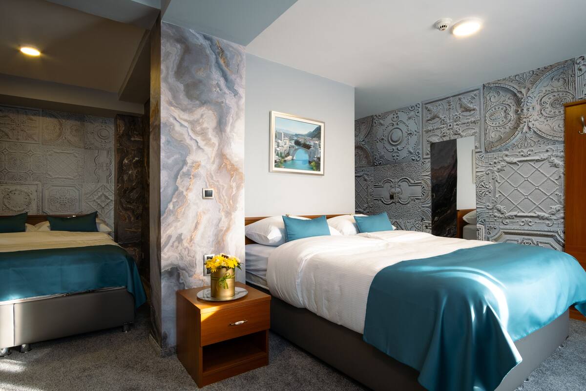 Ein modernes Hotelzimmer mit zwei Betten, Marmor-Wandverzierungen, einem gerahmten Stadtbild und einem Holznachttisch mit Blumen in einer Vase.