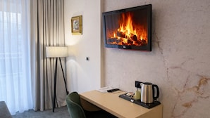 A hotel room with a desk, a chair, a flat-screen TV displaying a fireplace, a lamp, and a coffee pot. - Hotel Villa Harmony (Sarajevo)
