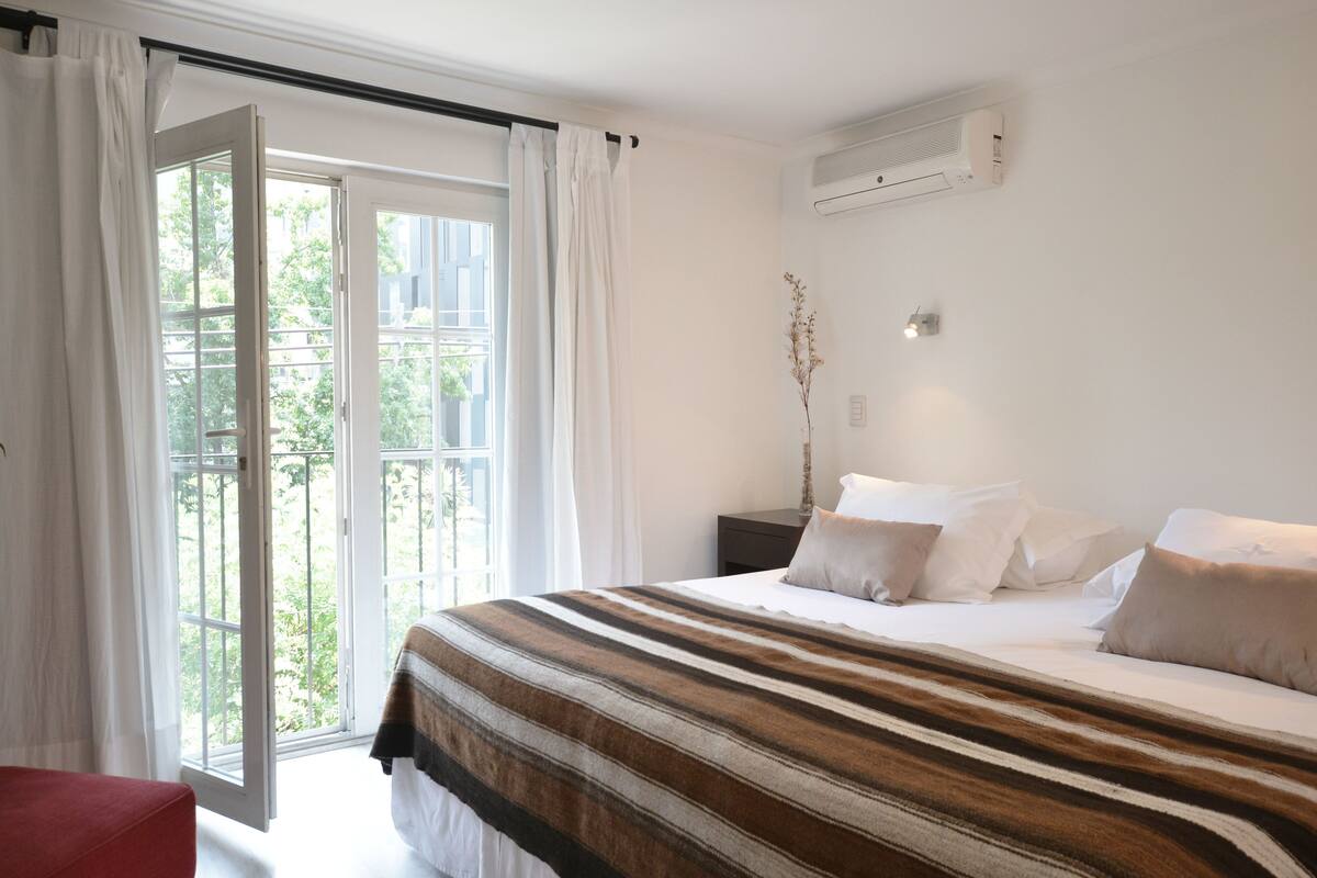 A bedroom with a large bed, a nightstand, a window with curtains, and an air conditioning unit.