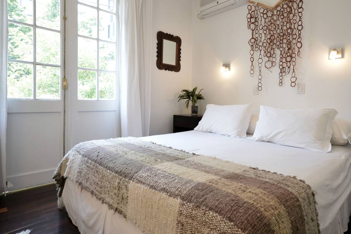 A neatly made bed with a striped blanket, a bedside table with a plant, a wall-mounted mirror, and a hanging light fixture.