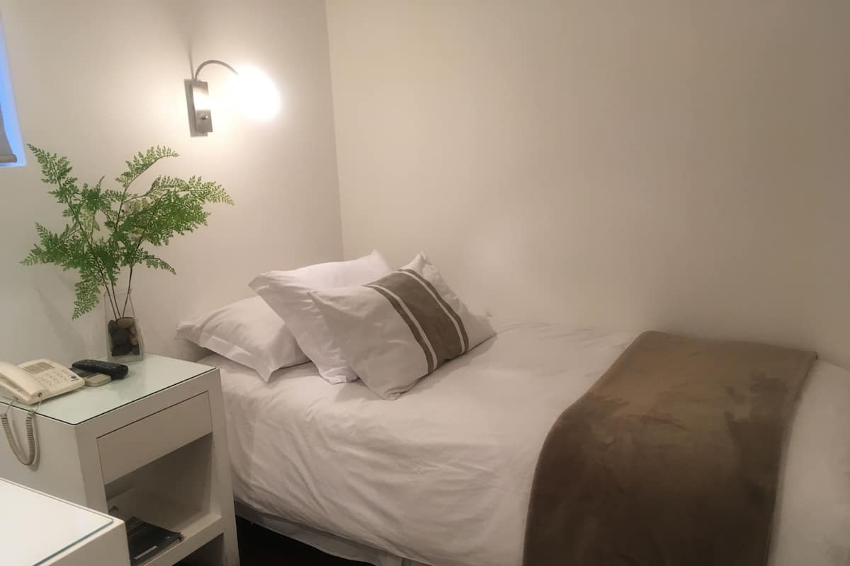 A neatly made bed with white bedding and a brown throw blanket, a bedside table with a phone and remote control, and a wall-mounted light fixture.