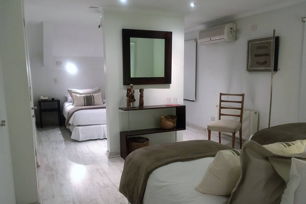 A bedroom with two beds, a chair, a shelf with decorative items, and an air conditioning unit.