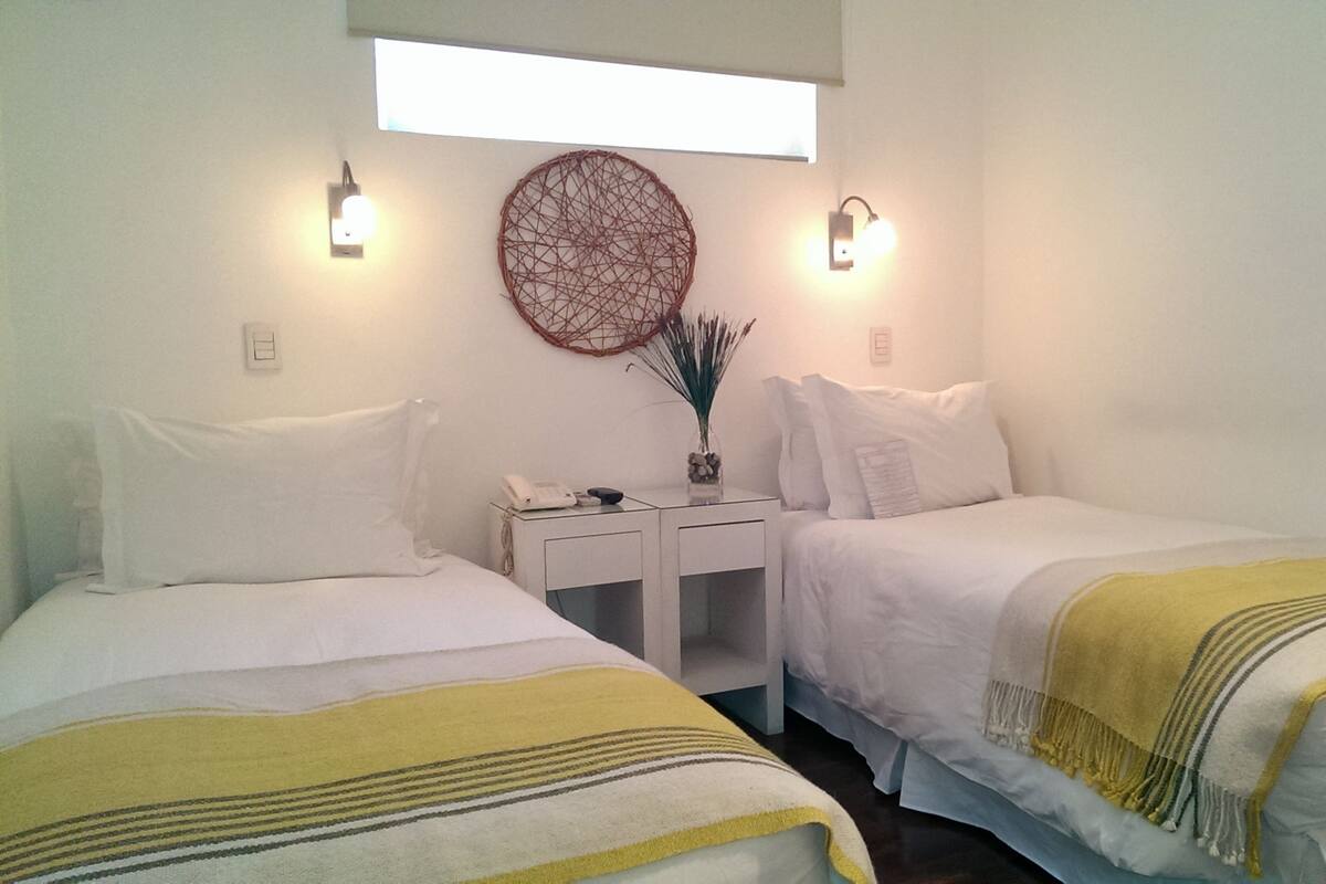 A hotel room with two beds, a small table with a phone, a decorative wall hanging, and wall-mounted lights.
